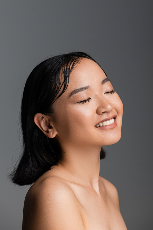 young asian woman with bare shoulders and nude makeup smiling with closed eyes isolated on grey