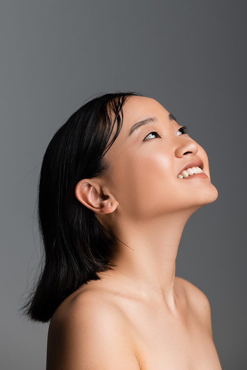 pleased asian woman with naked shoulders and nude makeup looking away isolated on grey