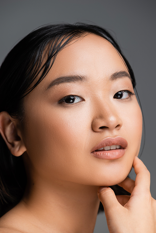 portrait of brunette asian woman with nude makeup looking at camera isolated on grey