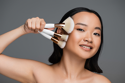 happy asian woman with bare shoulders and natural makeup holding different cosmetic brushes isolated on grey