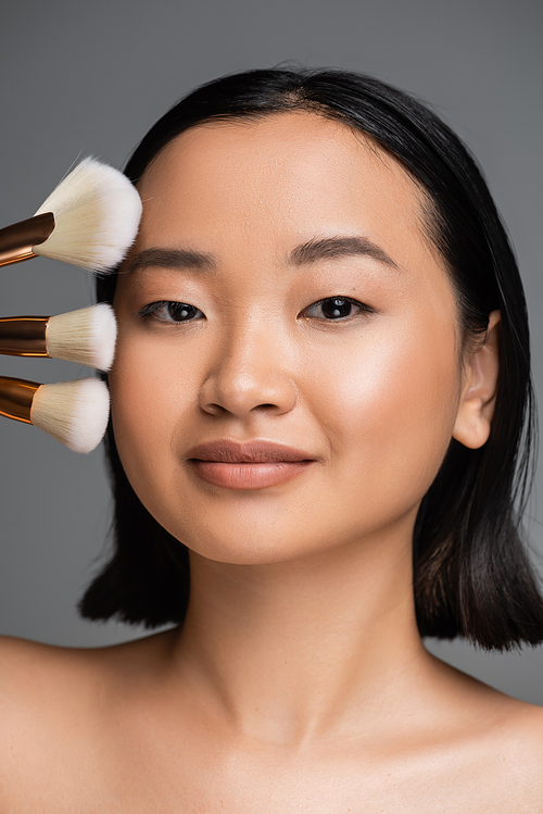 portrait of brunette asian woman with natural makeup looking at camera near different cosmetic brushes isolated on grey