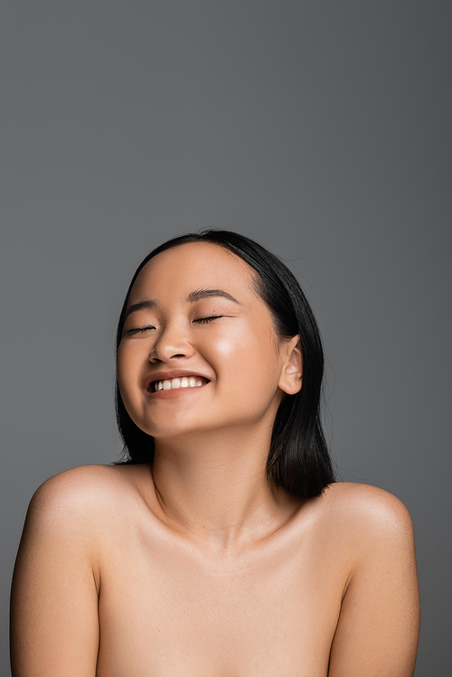 cheerful asian woman with bare shoulders and natural makeup smiling with closed eyes isolated on grey