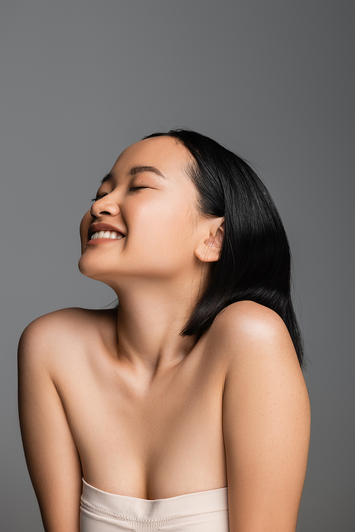 pleased asian woman with naked shoulders and closed eyes posing isolated on grey