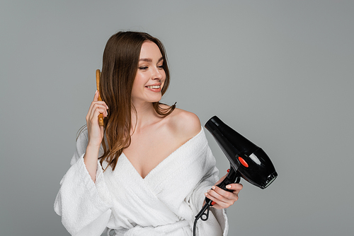 happy young woman with shiny hair using hair dryer and brushing hair isolated on grey