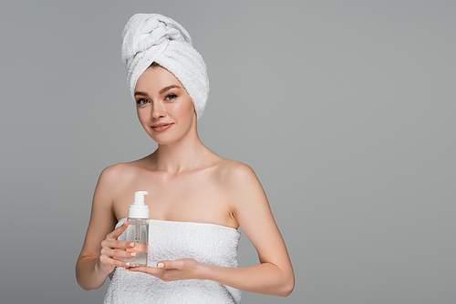 smiling young woman with bare shoulders and towel on head holding bottle with cleansing foam isolated on grey