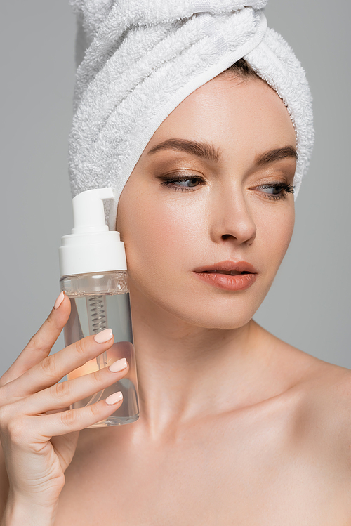 portrait of young woman with towel on head holding bottle with cleansing foam isolated on grey