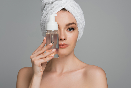 young woman with bare shoulders and towel on head holding bottle with cleansing foam near face isolated on grey