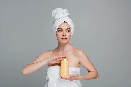 young woman with bare shoulders and towel on head holding bottle with shampoo isolated on grey