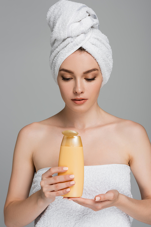 pretty woman with bare shoulders and towel on head holding bottle with shampoo isolated on grey