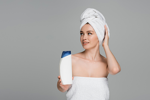 cheerful young woman with bare shoulders and towel on head holding bottle with shampoo isolated on grey