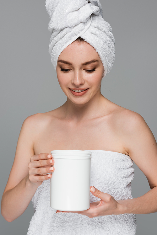 cheerful young woman with towel on head holding container with hair mask isolated on grey
