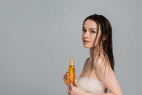 pretty young woman with wet hair and bare shoulders holding bottle with oil isolated on grey