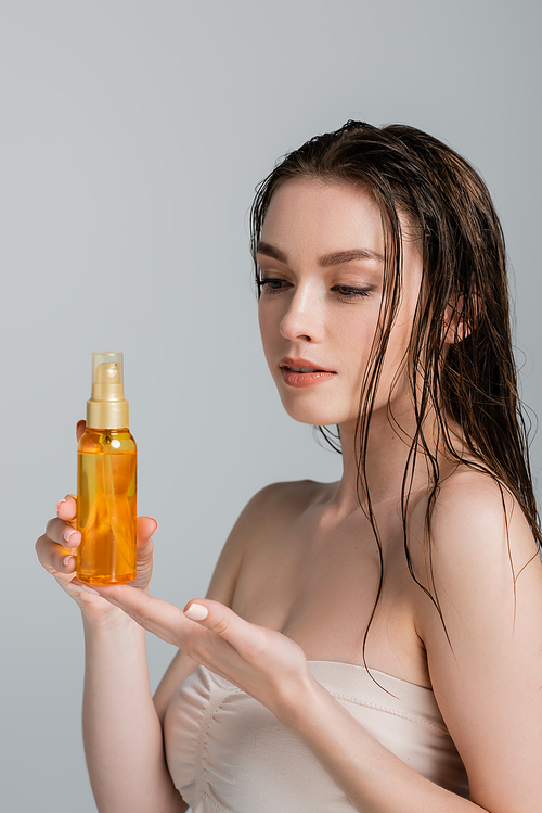 pretty young woman with bare shoulders looking at bottle with hair oil isolated on grey