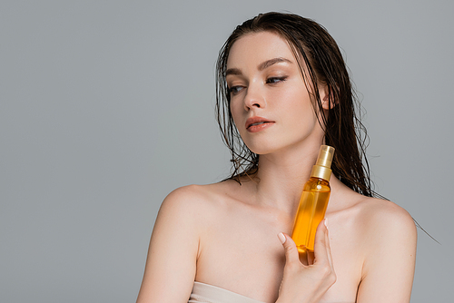 young woman with bare shoulders holding bottle with hair oil isolated on grey