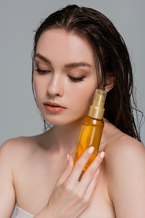 portrait of pretty young woman with bare shoulders holding bottle with hair oil isolated on grey