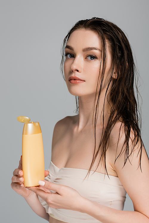 pretty young woman with wet hair and bare shoulders holding bottle with shampoo isolated on grey