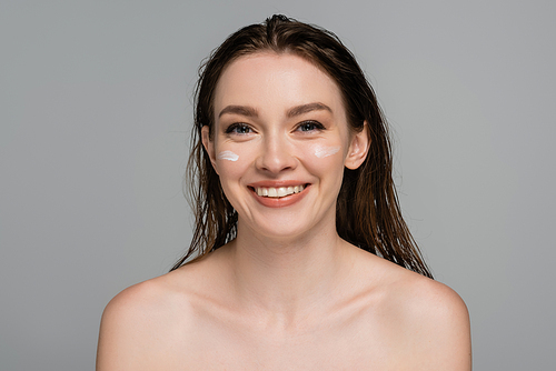 cheerful young woman with wet hair and moisturizing cream on cheeks isolated on grey