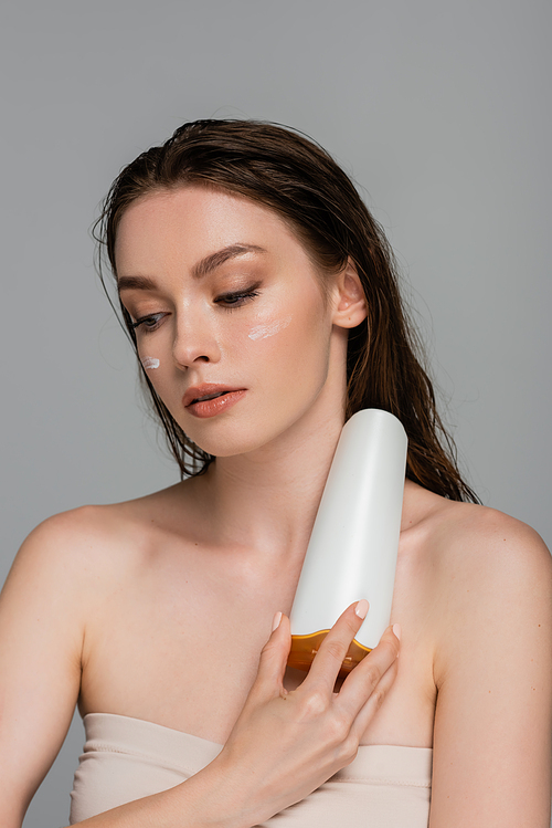 pretty young woman with wet hair and moisturizing cream on cheeks holding bottle with shampoo isolated on grey