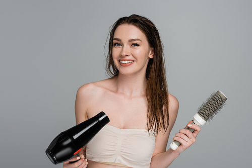 cheerful young woman with wet hair holding round hair brush and hair dryer isolated on grey
