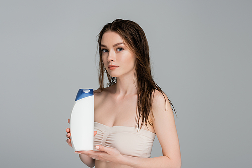 pretty woman with wet hair and bare shoulders holding shampoo bottle isolated on grey