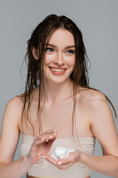 young and smiling woman with wet hair holding foam in hand isolated on grey