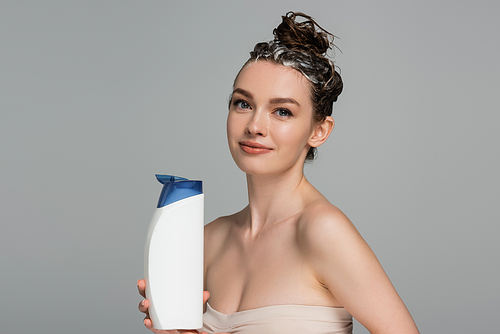 cheerful young woman with wet foamy hair holding bottle with shampoo isolated on grey