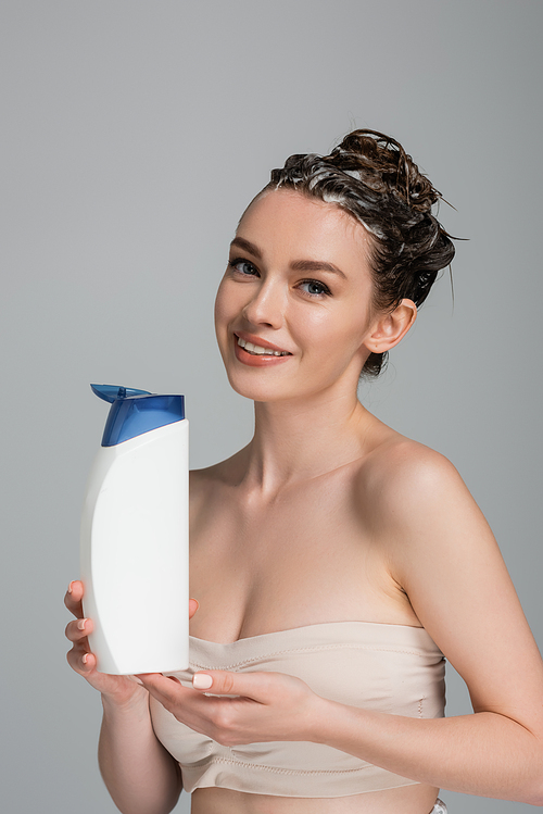 smiling young woman with wet foamy hair holding bottle with shampoo isolated on grey