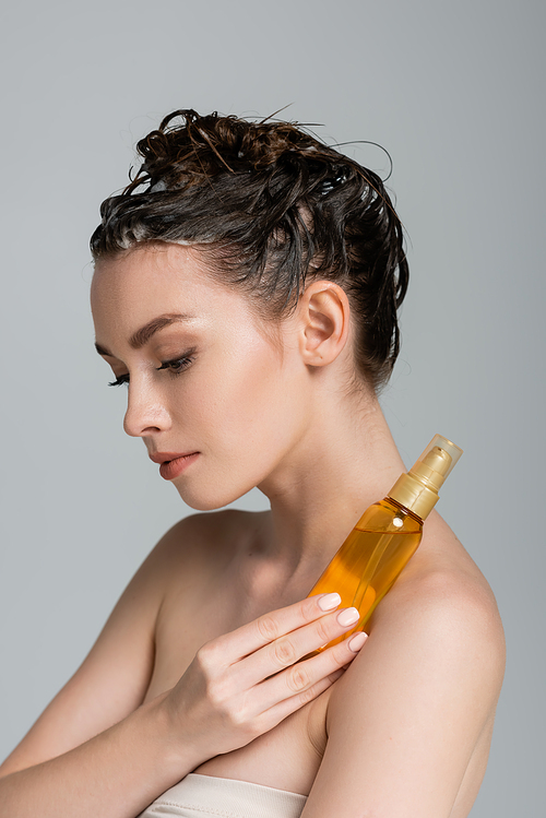 brunette young woman with wet hair holding bottle with treatment oil isolated on grey