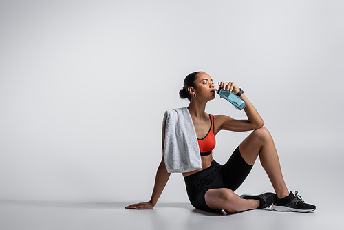 young african american woman in sports bra and bike shorts sitting with towel and drinking water on grey background