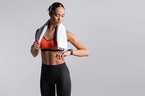 brunette african american sportswoman in red sports bra standing with towel and looking at fitness tracker isolated on grey
