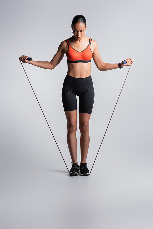 full length of sportive african american woman standing with skipping rope on grey