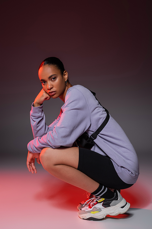 full length of african american woman in bike shorts and sweatshirt sitting on grey with red light