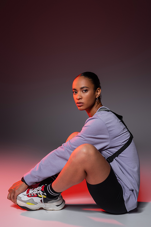 full length of young african american woman in bike shorts and sweatshirt sitting on grey with red light