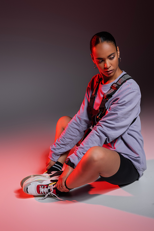 full length of stylish african american woman in bike shorts and sweatshirt sitting on grey with red light