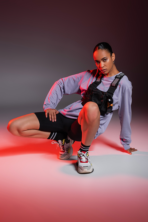 full length of african american woman in bike shorts and sweatshirt sitting while posing on grey with red light