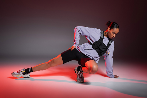 full length of african american woman in bike shorts and sweatshirt stretching leg on grey background with red light