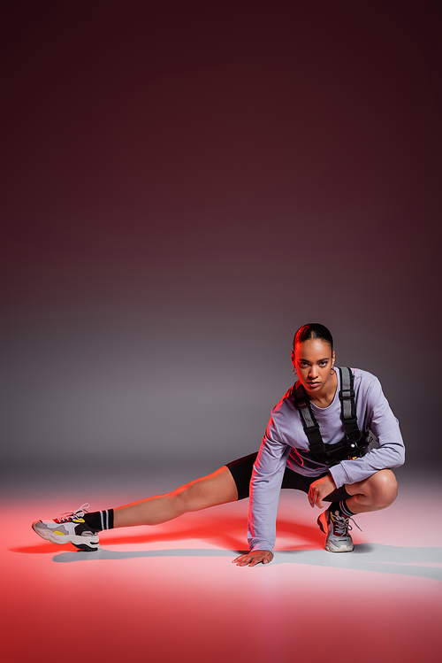 full length of sporty african american woman in bike shorts and sweatshirt stretching leg on grey with red light