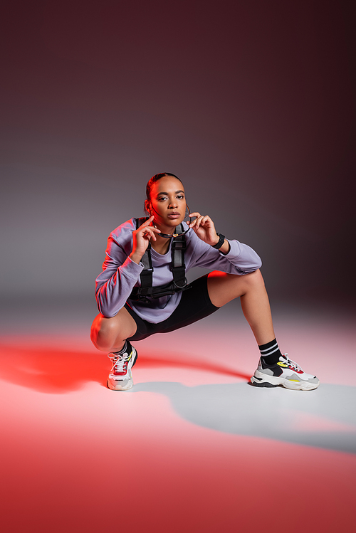 full length of african american woman in bike shorts and sweatshirt sitting while wearing sunglasses on grey background with red light