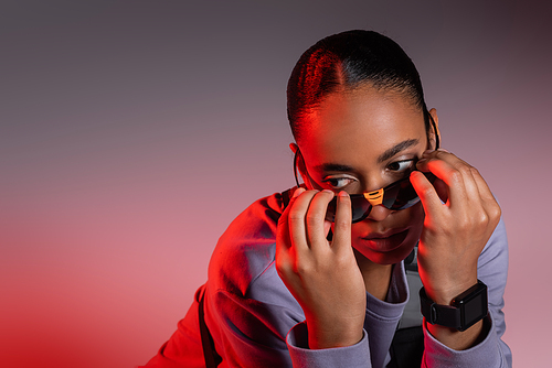african american woman in sweatshirt wearing stylish sunglasses on grey background with red light