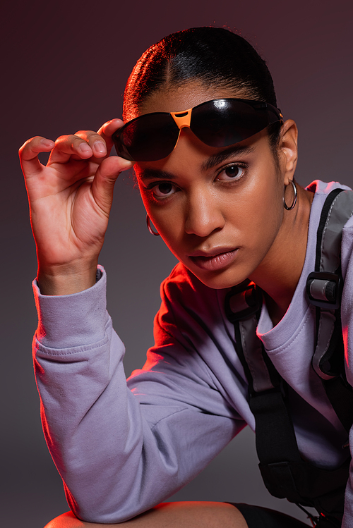 african american woman in sweatshirt adjusting stylish sunglasses on grey with red light