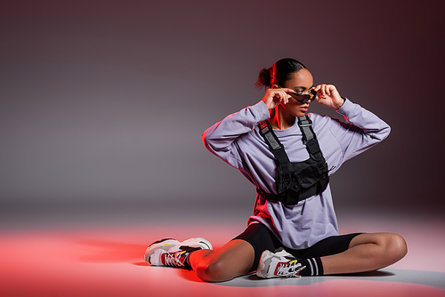 full length of african american woman in bike shorts and sweatshirt wearing sunglasses on grey background with red light