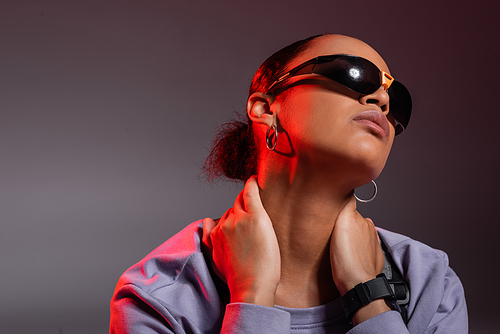 portrait of african american woman in sweatshirt and trendy sunglasses touching neck on grey with red light