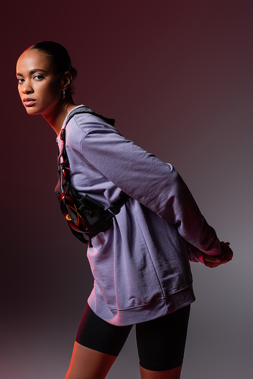 young african american woman in sweatshirt and vest looking at camera on grey and purple
