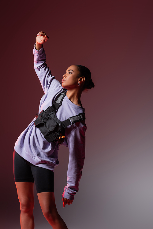 young african american woman in sweatshirt and bike shorts posing with raised hand on grey and purple