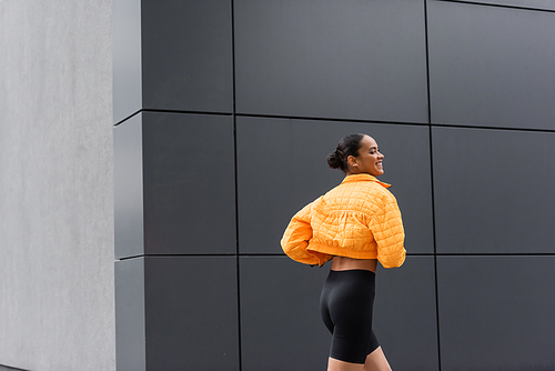 cheerful african american sportswoman in bike shorts and yellow puffer jacket exercising outside