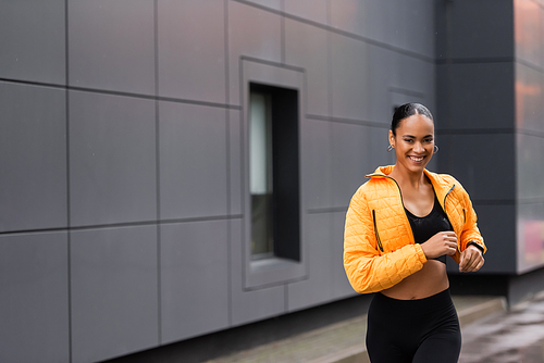 positive african american sportswoman in yellow puffer jacket jogging outside