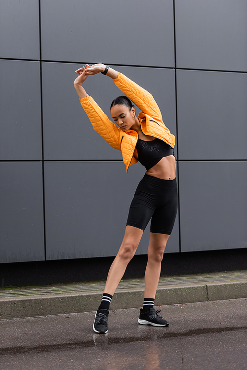 full length of brunette african american sportswoman in bike shorts and puffer jacket warming up outside