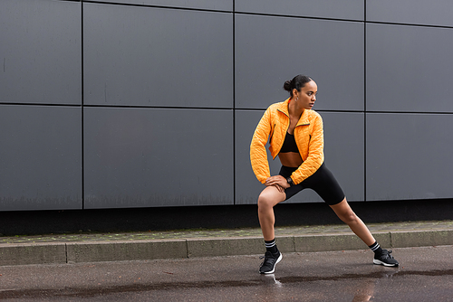 full length of brunette african american sportswoman in bike shorts and yellow puffer jacket exercising outside