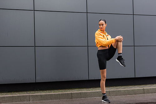 full length of brunette african american sportswoman in bike shorts and yellow puffer jacket working out near building
