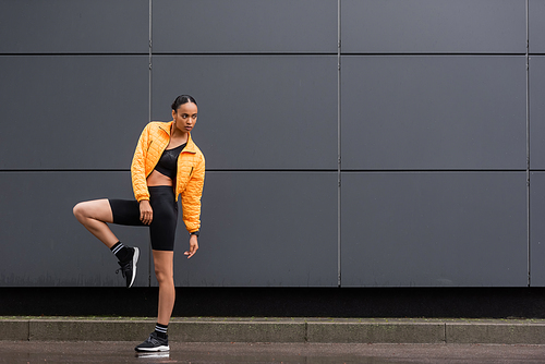 full length of brunette african american sportswoman in bike shorts and yellow puffer jacket posing outside